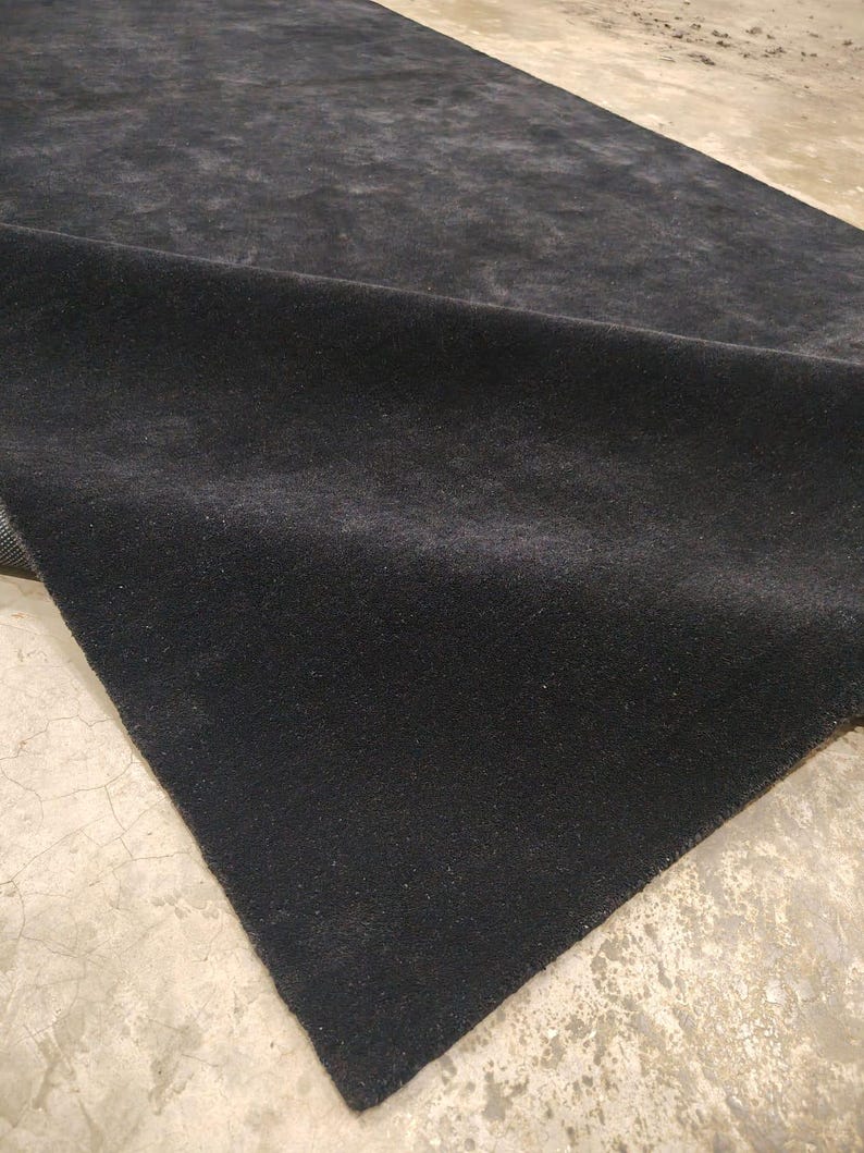 May include: A rectangular area rug with a solid black, dense, and short-pile surface. The rug is shown partially folded over, revealing the dark texture of the material and its straight, finished edges against a light-colored floor.