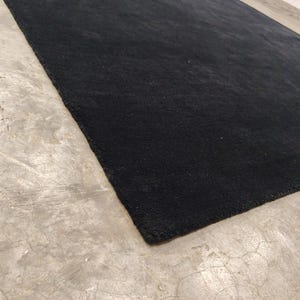 May include: A large, rectangular area rug featuring a solid, deep black color with a dense, short-pile texture. The rug is laid out flat, showcasing its clean, straight edges and uniform, dark surface across the entire visible area.