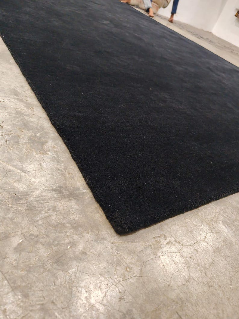 May include: A large, rectangular area rug featuring a solid, deep black color with a dense, short-pile texture. The rug is laid out flat, showcasing its clean, straight edges and uniform, dark surface across the entire visible area.