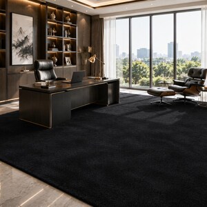 May include: A large, solid black rectangular area rug with a dense, low-pile texture. The rug covers a significant portion of the floor in a home office, extending from the base of a dark wood desk to the seating area with a lounge chair.
