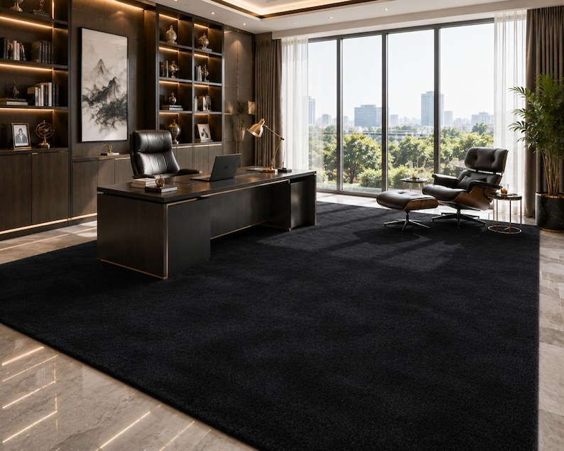 May include: A large, solid black rectangular area rug with a dense, low-pile texture. The rug covers a significant portion of the floor in a home office, extending from the base of a dark wood desk to the seating area with a lounge chair.