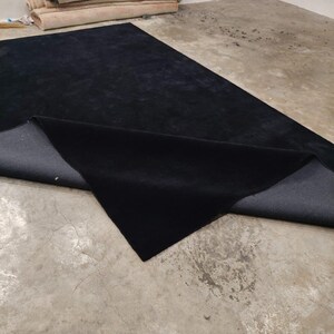 May include: A large rectangular area rug with a solid black pile surface and a dark gray woven backing. One corner of the rug is folded over to reveal the contrasting dark gray underside, showcasing the texture of the material.