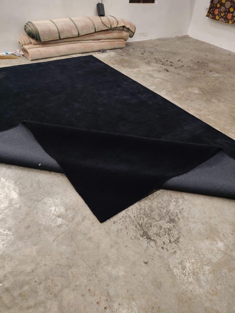 May include: A large rectangular area rug with a solid black pile surface and a dark gray woven backing. One corner of the rug is folded over to reveal the contrasting dark gray underside, showcasing the texture of the material.