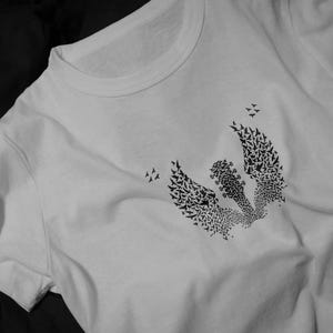 Op de afbeelding: Wit t-shirt met een zwart grafisch ontwerp. Het ontwerp toont een gitaar met vleugels, gevormd door vele kleine vogels. Het ontwerp is centraal geplaatst, met meer vogels eromheen.