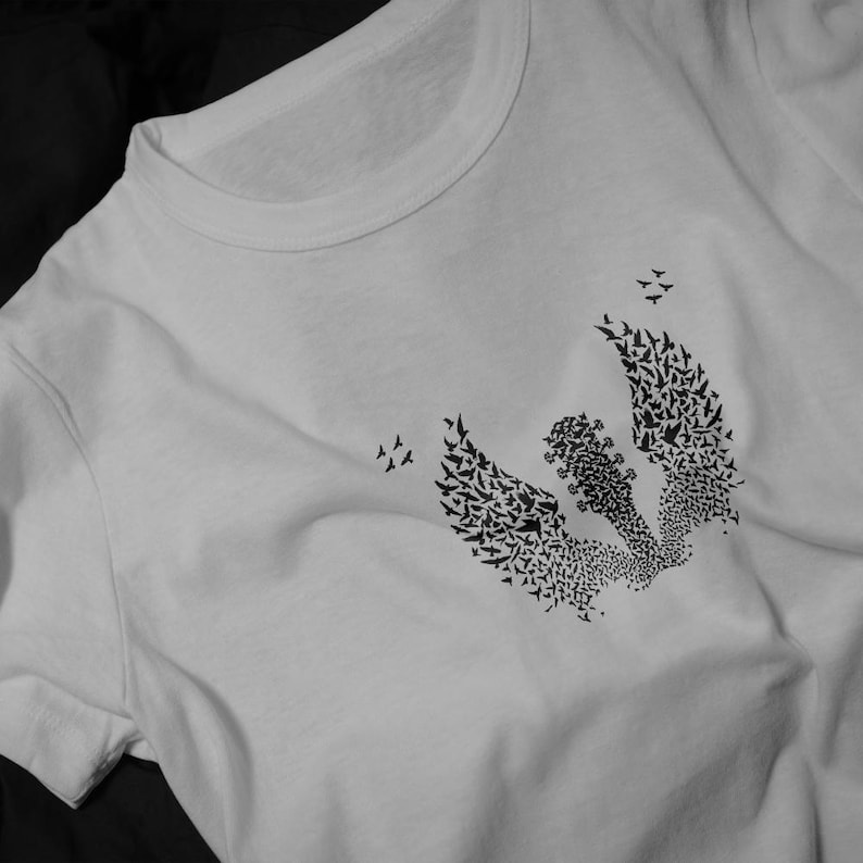 Op de afbeelding: Wit t-shirt met een zwart grafisch ontwerp. Het ontwerp toont een gitaar met vleugels, gevormd door vele kleine vogels. Het ontwerp is centraal geplaatst, met meer vogels eromheen.