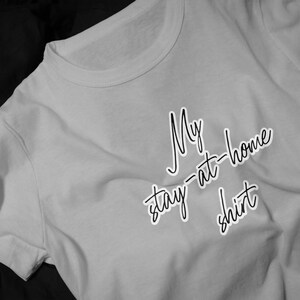 May include: A light gray short-sleeved t-shirt featuring the black cursive text My stay-at-home shirt printed on the front. The garment has a classic crew neckline and a simple, casual design with a soft, folded fabric texture visible throughout.