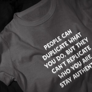 以下が含まれることがあります： ダークグレーのクルーネックTシャツ。白い文字で「PEOPLE CAN DUPLICATE WHAT YOU DO, BUT THEY CAN'T REPLICATE WHO YOU ARE STAY AUTHENTIC」とプリントされています。文字にはかすかなドロップシャドウ効果があり、生地の上で重なり合っているような見た目です。