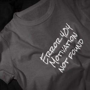 Pode incluir: Uma t-shirt cinzento-escura de gola redonda com texto branco impresso num estilo inclinado e desenhado à mão. O texto diz "ERROR 404 MOTIVATION NOT FOUND" com um efeito de sombra subtil, criando uma aparência em camadas no tecido.