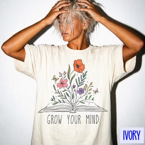 Puede incluir: Camiseta de color marfil con un diseño floral que presenta un libro abierto y el texto "GROW YOUR MIND". La ilustración incluye amapolas naranjas, flores rosas y moradas, y mariposas. La camiseta es de un color suave y neutro.