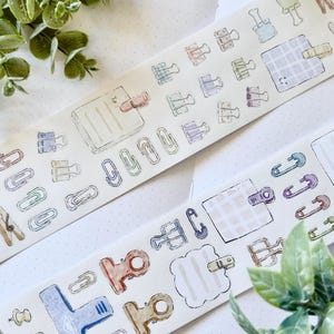 Peut inclure: Deux rouleaux de ruban adhésif décoratif avec des illustrations à l'aquarelle de fournitures de bureau telles que des trombones, des pinces à dessin et des punaises. Le ruban a un fond crème avec un motif à pois et est orné de verdure.