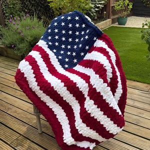 Könnte beinhalten: Eine handgestrickte Decke im Design der amerikanischen Flagge. Sie hat ein marineblaues Feld mit weißen Sternen und abwechselnd roten und weißen Streifen. Die Decke besteht aus einem weichen, plüschigen Material.