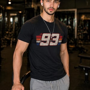 May include: A black short-sleeved t-shirt featuring the number 93 in a distressed white font, centered over horizontal red, white, and blue stripes. The shirt has a crew neckline and is paired with grey drawstring sweatpants.