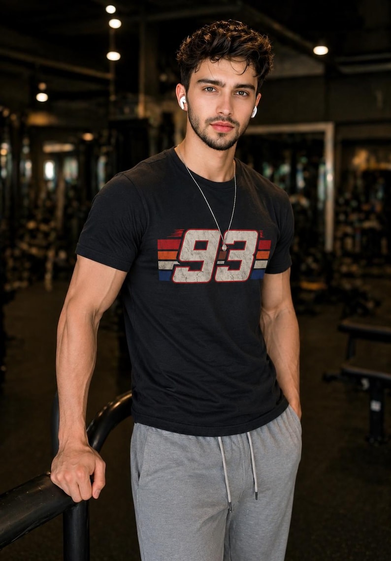 May include: A black short-sleeved t-shirt featuring the number 93 in a distressed white font, centered over horizontal red, white, and blue stripes. The shirt has a crew neckline and is paired with grey drawstring sweatpants.