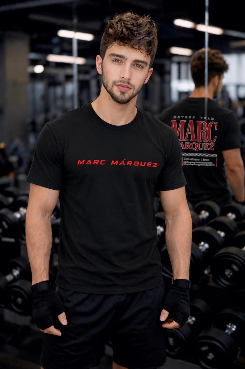 May include: A black short-sleeved crew neck t-shirt featuring the text MARC M&Aacute;RQUEZ in red lettering on the chest. The back of the shirt displays the text MOTO GP TEAM MARC M&Aacute;RQUEZ in red and white print. The shirt is worn with black fingerless gloves.