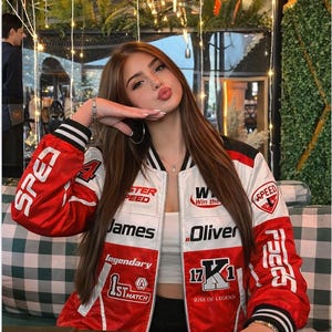 May include: A red, white, and black racing jacket with the words "SPEED", "James", and "Oliver" printed on it. The jacket has a white collar and black and white striped cuffs. The jacket is worn over a white top.