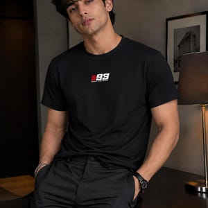 May include: A black short-sleeved crew neck t-shirt featuring a small graphic on the chest with the number 93 in white, red vertical stripes, and the text marc marquez printed in white lettering below the number on a plain black background.
