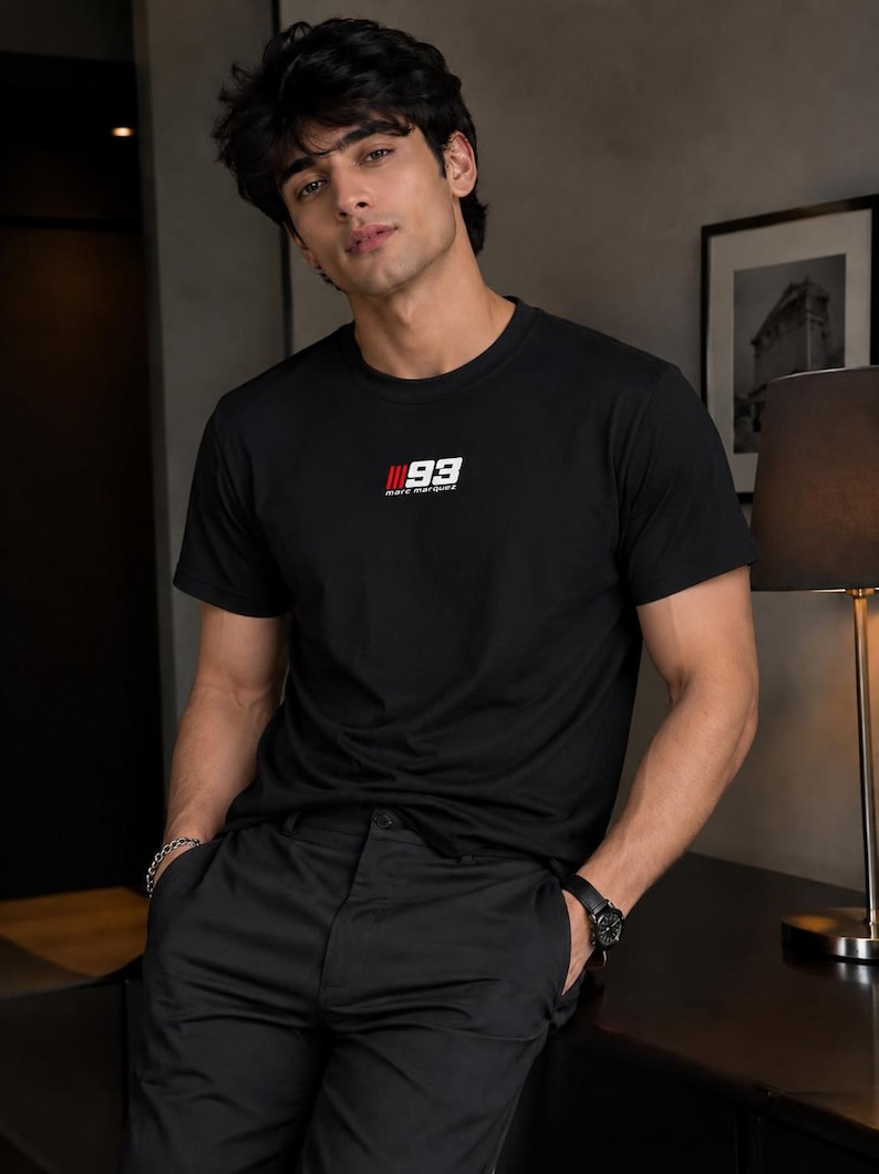 May include: A black short-sleeved crew neck t-shirt featuring a small graphic on the chest with the number 93 in white, red vertical stripes, and the text marc marquez printed in white lettering below the number on a plain black background.