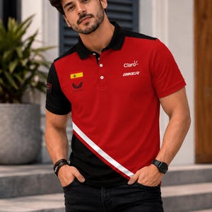 Op de afbeelding: Een rood en zwart poloshirt met korte mouwen, voorzien van een witte diagonale streep, een zwarte kraag met knopen en de tekst Claro en BIKER op de borst. Het shirt toont ook een kleine Spaanse vlag en een logo aan de voorzijde.