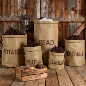 May include: A rustic kitchen storage set featuring five brown paper sacks labeled "Potatoes," "Onions," "Bread," "Vegetables," and "Garlic." Each sack has a dark brown drawstring closure and a decorative stitched seam. A wooden crate filled with produce sits in front.