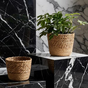 Puede incluir: Dos macetas tejidas de color marrón claro. Una está vacía, la otra contiene una planta verde. Las macetas se exhiben sobre una superficie de mármol blanco y negro, creando un contraste de texturas y colores. Las macetas son de fibra natural.