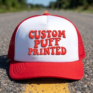 Può includere: Un cappellino trucker rosso e bianco con un pannello frontale bianco che riporta la scritta rossa CUSTOM PUFF PRINTED in un carattere grassetto e arrotondato. Il cappello presenta una visiera rossa e pannelli laterali e posteriori in rete.