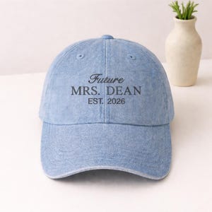 Puede incluir: Gorra de béisbol de mezclilla azul claro con el texto "Future MRS. DEAN EST. 2026" en escritura negra. La gorra tiene una visera curva y un botón en la parte superior. Un jarrón decorativo con vegetación está en el fondo.