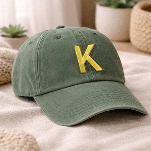 May include: A washed olive green baseball cap featuring a prominent, embroidered yellow letter K on the front panel. The cap has a structured crown, a curved brim with visible stitching, and a button at the top center of the six-panel design.
