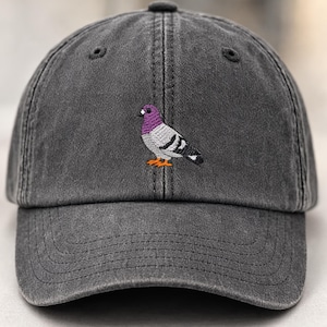以下が含まれることがあります： 色あせた仕上がりのチャコールグレーの野球帽。紫、グレー、黒、オレンジのアクセントが施された、刺繍されたハトのデザインが特徴です。カーブしたつばと調節可能なストラップが付いています。