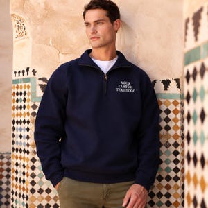 May include: A navy blue quarter-zip sweatshirt featuring a high collar, long sleeves, and ribbed cuffs. The chest area displays the white embroidered text "YOUR CUSTOM TEXT/LOGO". The garment is worn over a plain white crew neck t-shirt.