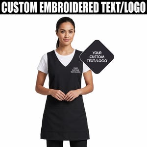 Op de afbeelding: Een zwarte schort met een V-hals, gedragen over een wit t-shirt. De schort heeft een zak en is voorzien van de tekst "YOUR CUSTOM TEXT/LOGO". De afbeelding bevat ook een zwarte ruitvormige afbeelding met de tekst "YOUR CUSTOM TEXT/LOGO". De tekst "CUSTOM EMBROIDERED TEXT/LOGO" staat bovenaan.