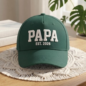 May include: A forest green baseball cap featuring the word PAPA in large, white, raised block letters across the front, with the text EST. 2026 printed in smaller white letters directly underneath the main word on the structured crown.