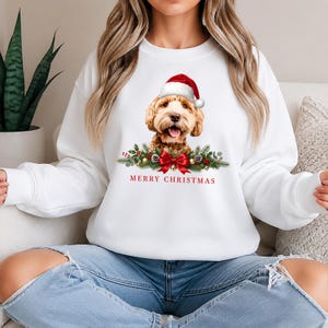 May include: A white crewneck sweatshirt featuring a graphic of a golden-coloured dog wearing a red and white Santa hat. Below the dog is a festive arrangement of pine branches, red berries, and a red bow, with the text MERRY CHRISTMAS in red.