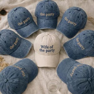 May include: A collection of eight baseball caps arranged in a circle. Seven caps are made of blue denim with the text 'the party' embroidered in gold. One central cap is made of off-white fabric with the text 'Wife of the party' in navy blue.