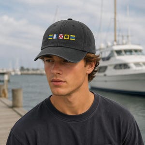 May include: A dark grey baseball cap featuring embroidered nautical flag symbols. The cap has a curved peak and a classic six-panel design. A boat and a dock are visible in the background.