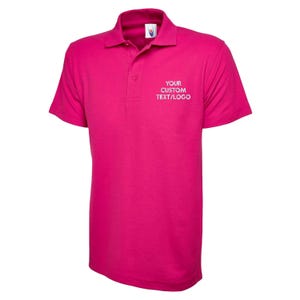 May include: A bright magenta polo shirt with short sleeves and a collared neckline. The shirt has a three-button placket and the text "YOUR CUSTOM TEXT/LOGO" in white on the chest. The shirt is made of a smooth fabric.