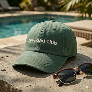Puede incluir: Gorra de béisbol verde con el texto blanco "cool dad club". La gorra está sobre una superficie de piedra junto a unas gafas de sol carey.