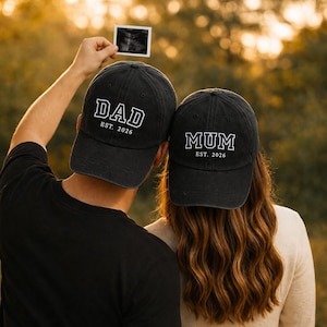 May include: Two black baseball caps with white lettering that reads "DAD EST. 2026" and "MUM EST. 2026". The image shows the backs of two people, one holding a sonogram. The background is blurred with warm tones.