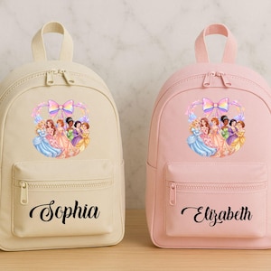 May include: Two small backpacks, one cream and one pink, each with a princess graphic and a name. The cream backpack has "Sophia" in script, while the pink one says "Elizabeth". Both have a front pocket and top handle.