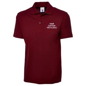 May include: A maroon polo shirt with short sleeves and a collar. The shirt features a three-button placket and the text "YOUR CUSTOM TEXT/LOGO" in white on the chest. The shirt is made of a soft, breathable fabric.