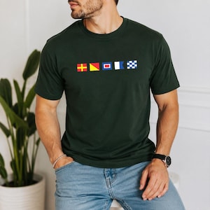 Op de afbeelding: Een donker bosgroen T-shirt met korte mouwen en een ronde hals, voorzien van een horizontale rij met vijf kleurrijke nautische signaalvlaggen op het midden van de borst. De vlaggen hebben geometrische patronen in rood, geel, blauw en wit.