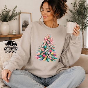 Può includere: Una felpa beige a girocollo con una grafica colorata di un albero di Natale stilizzato decorato con ornamenti rosa, turchesi e dorati, dettagli leopardati e un grande fiocco leopardato rosa sulla parte superiore.