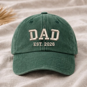 Puede incluir: Gorra de béisbol verde con la palabra "DAD" bordada en color beige claro. Debajo, el texto "EST. 2026". La gorra tiene una visera curva y un botón en la parte superior.