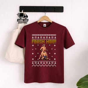 May include: A maroon short-sleeve t-shirt featuring a graphic print with the text "FINISH HIM!!" above two figures wearing Santa hats in a fighting stance, surrounded by a white holiday-themed pattern of snowflakes and pine trees.