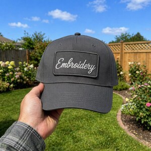 Könnte beinhalten: Eine anthrazitfarbene Baseballkappe mit gebogenem Schirm. Die Kappe hat einen rechteckigen Aufnäher mit dem Wort "Embroidery" in weißer Schrift. Die Kappe wird vor einem Hintergrund aus grünem Gras und blauem Himmel gehalten.
