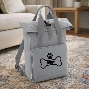 May include: A gray backpack with a roll-top closure, top handle, and adjustable shoulder straps. The front pocket features a black paw print and bone design with the name "RUBY". The bag has a buckle closure and a zippered pocket.