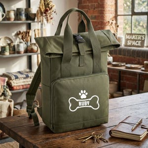 May include: Olive green backpack with a roll-top closure and a front pocket. The pocket features a white graphic of a dog bone with the name "RUBY". The bag has adjustable straps and a top handle. A notebook, pen, and keys are on the wooden surface.