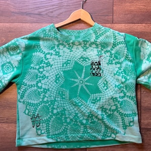 May include: Green short-sleeved t-shirt featuring a large white lace-like design. The shirt has a crew neck and a small juice box graphic with the text "HOB EL FSH CSS". The initials "AA JJ" are also visible.