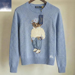 May include: Light blue knit sweater featuring a bear graphic wearing a hat, sweater, and jeans. The sweater has ribbed cuffs and hem, with the letters "RL" on the lower right. A tag with the brand name is attached.