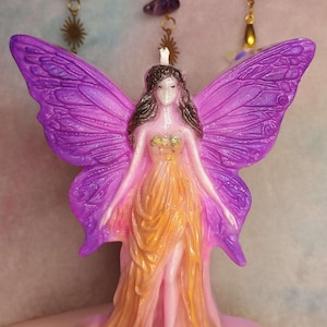 May include: A wax candle shaped like a fairy with large, textured purple wings. The figure has dark hair and wears a flowing, layered gown that transitions from pink to golden yellow. The bodice features gold accents, and a small white wick sits atop the head.