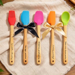 May include: A set of five kitchen utensils with bamboo handles and silicone heads in red, blue, pink, orange, and green. The handles feature engraved text including Life is better with frosting, Bake happy, spread heart, Whisk dreams, spread happiness, Create beauty, spread delight, and Make every dessert perfect.